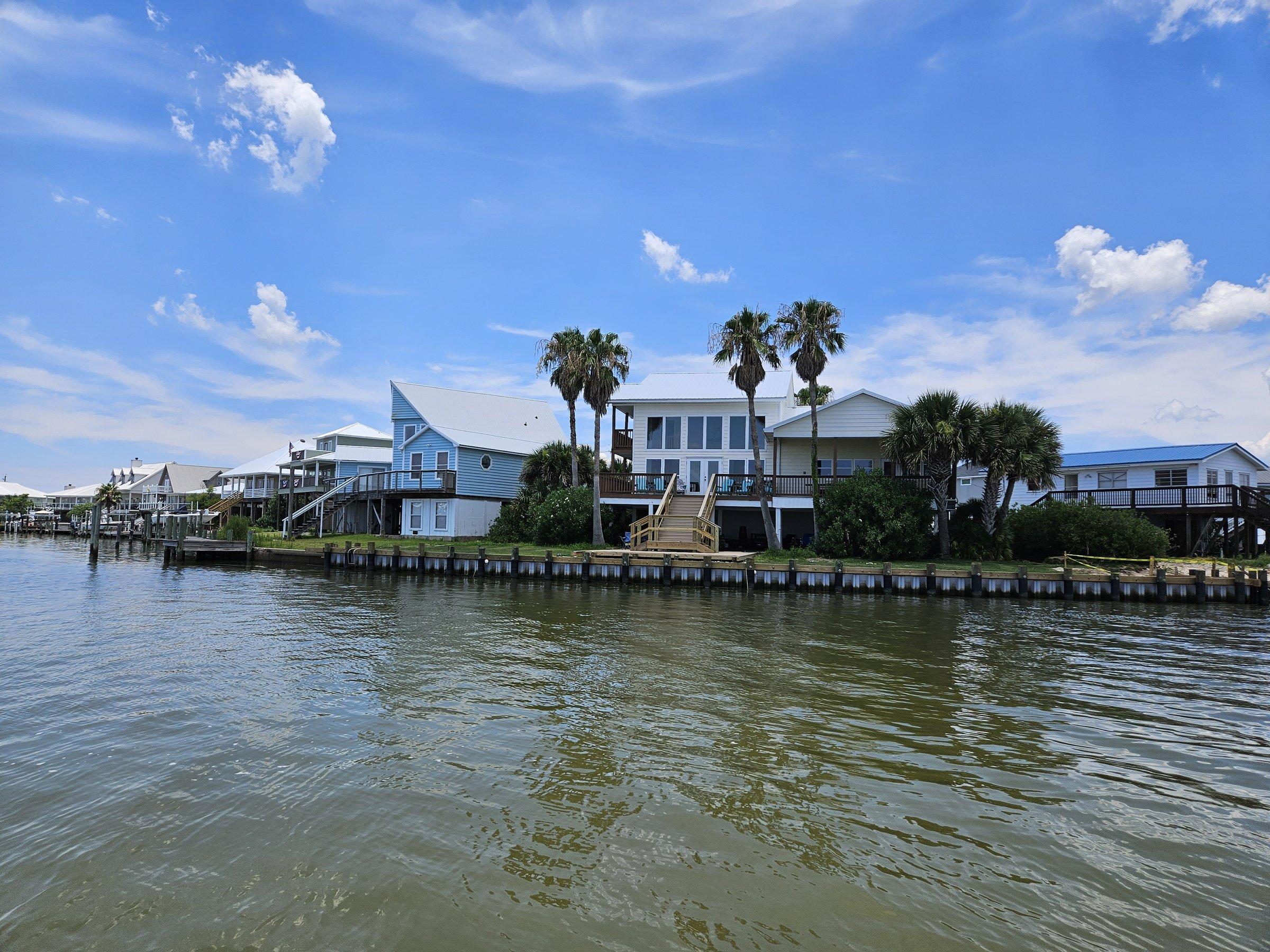 Elevated Gulf Coast homes with metal roofs and palm trees line a quiet canal under a blue sky.